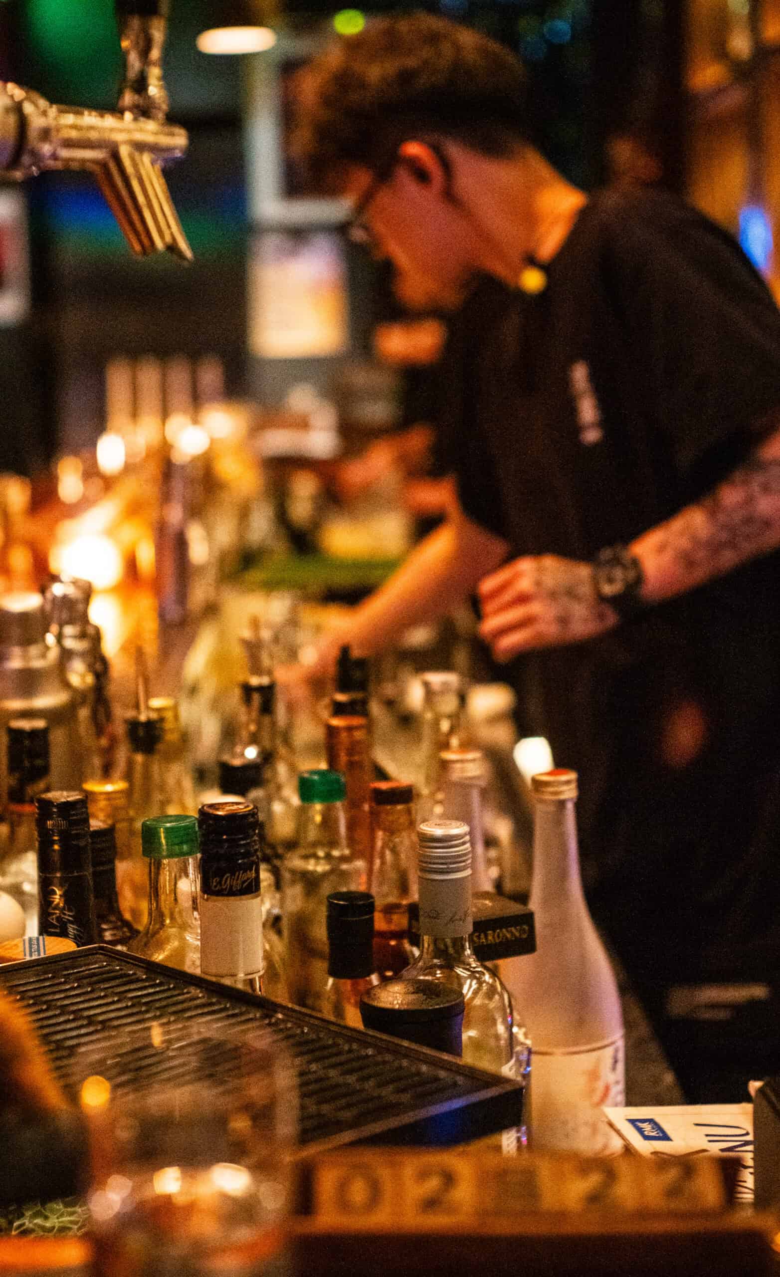 Man at bar, serving drinks, bottles, taps, tattoos, light night time