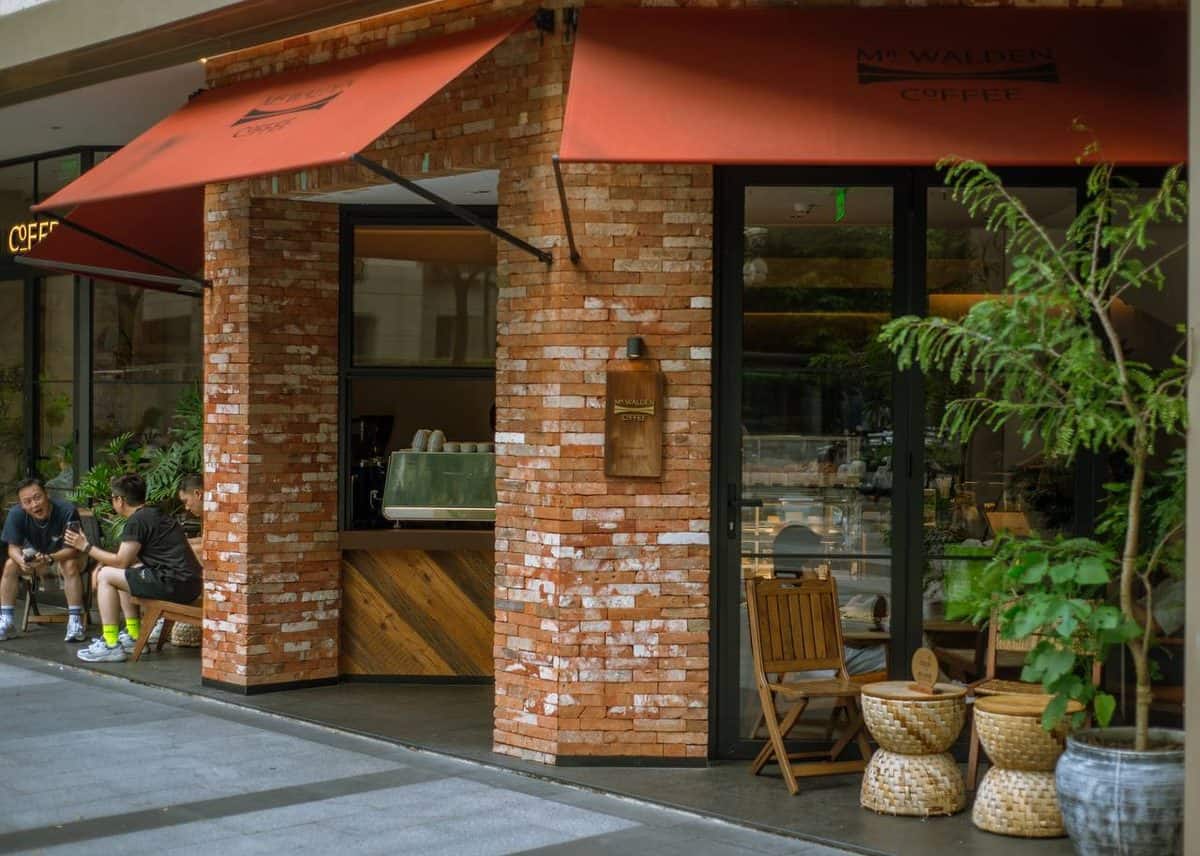 Walden's coffee exterior has red brick columns, red awnings, a yellow COFFEE sign, and three people on a bench.