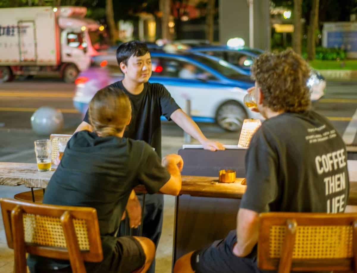 Three men at an outdoor table at Lotiko at night, one drinking beer while traffic passes behind.