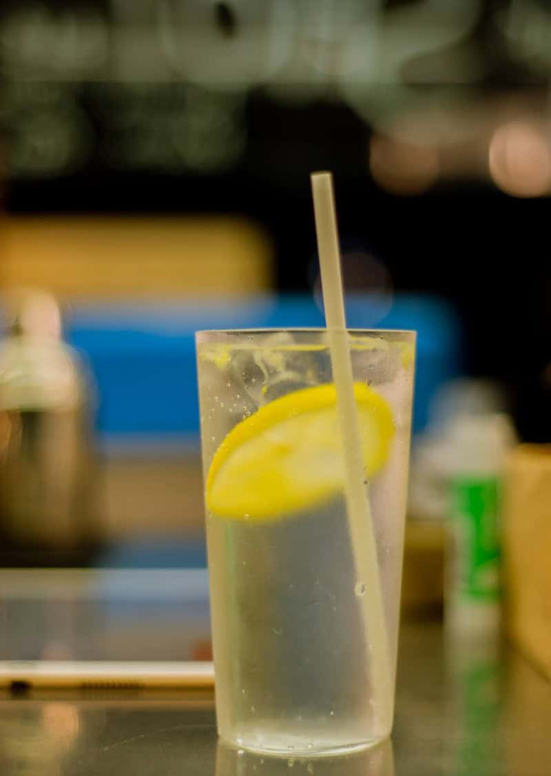 Tall glass of pale beverage with lemon and straw at Lotiko, blurred bar background behind.