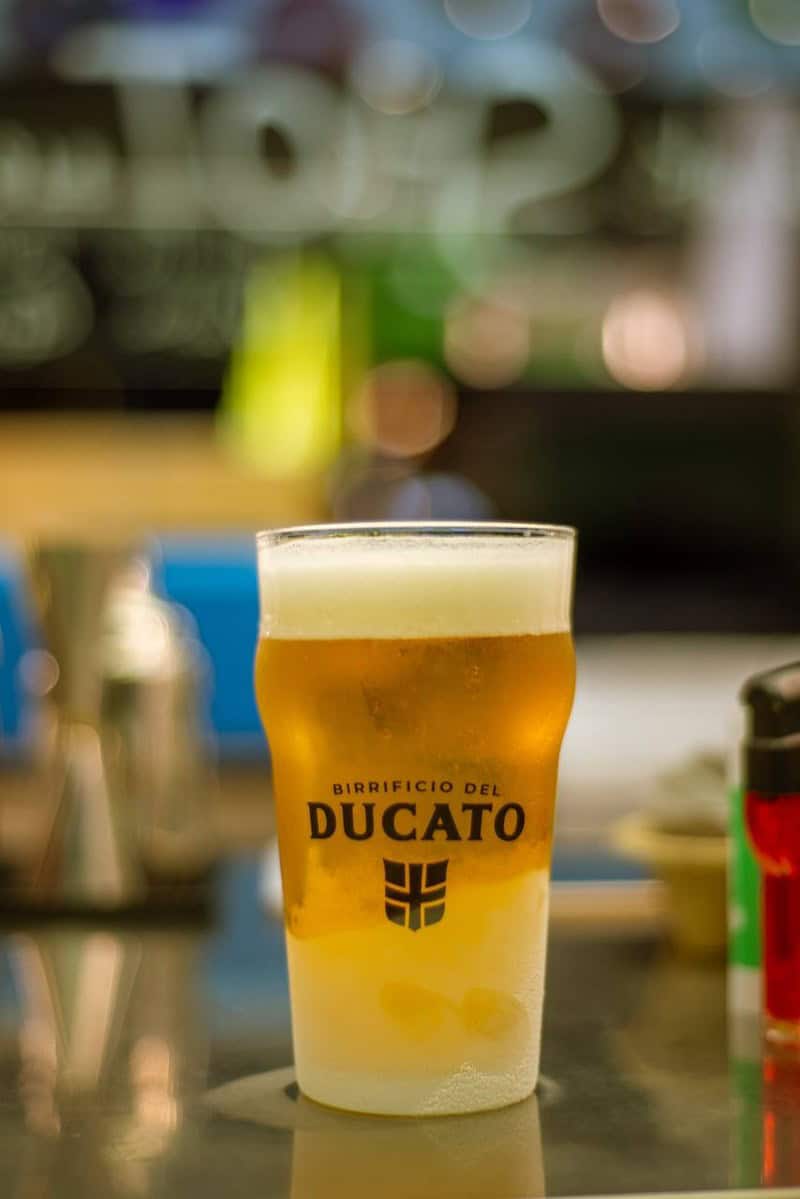 Pint glass printed with Birrificio del Ducato and shield-like logo holds amber beer with white foam at Lotiko.
