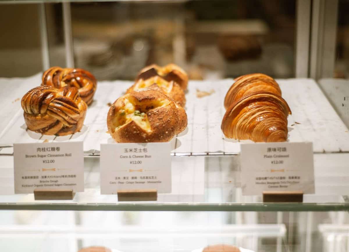 Cinnamon rolls, corn and cheese buns and croissants behind glass at Walden's coffee, with bilingual price signs.