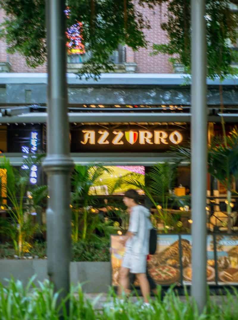 Pedestrian in a dark cap walks past Coco Park, framed by poles, plants, and an illuminated AZZURRO sign — thirstyguide.com