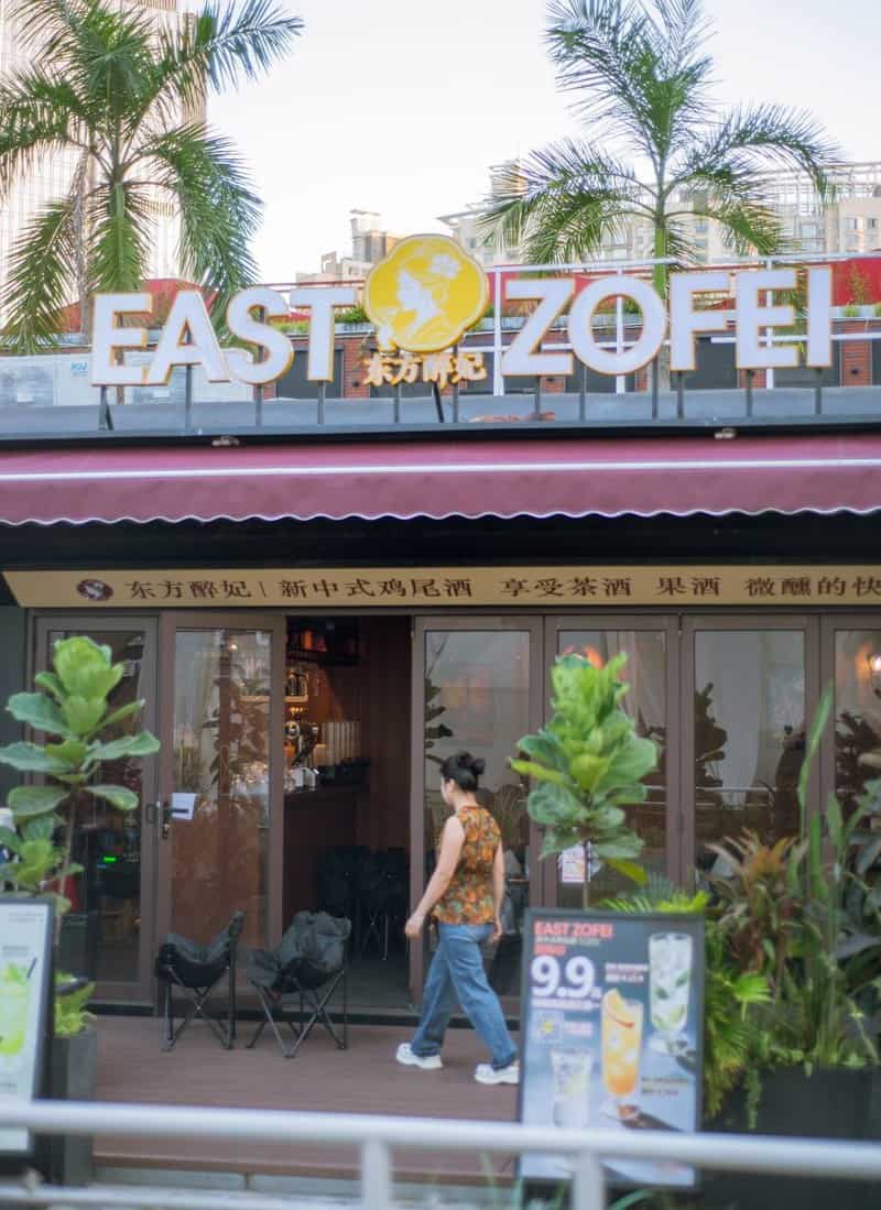A woman walks toward the Coco Park entrance past potted plants and palm trees — thirstyguide.com