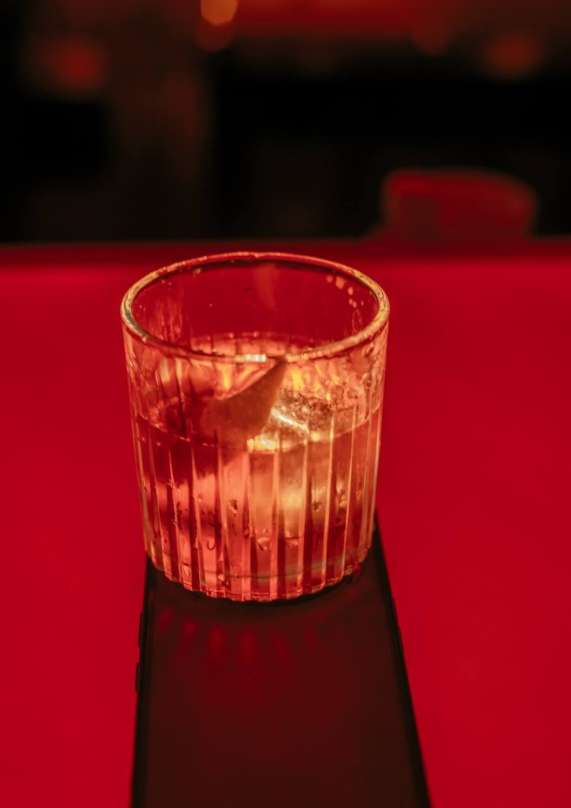A ribbed glass with clear liquid, ice, and citrus peel on a bright red surface at Scillia and Solo bar — thirstyguide.com